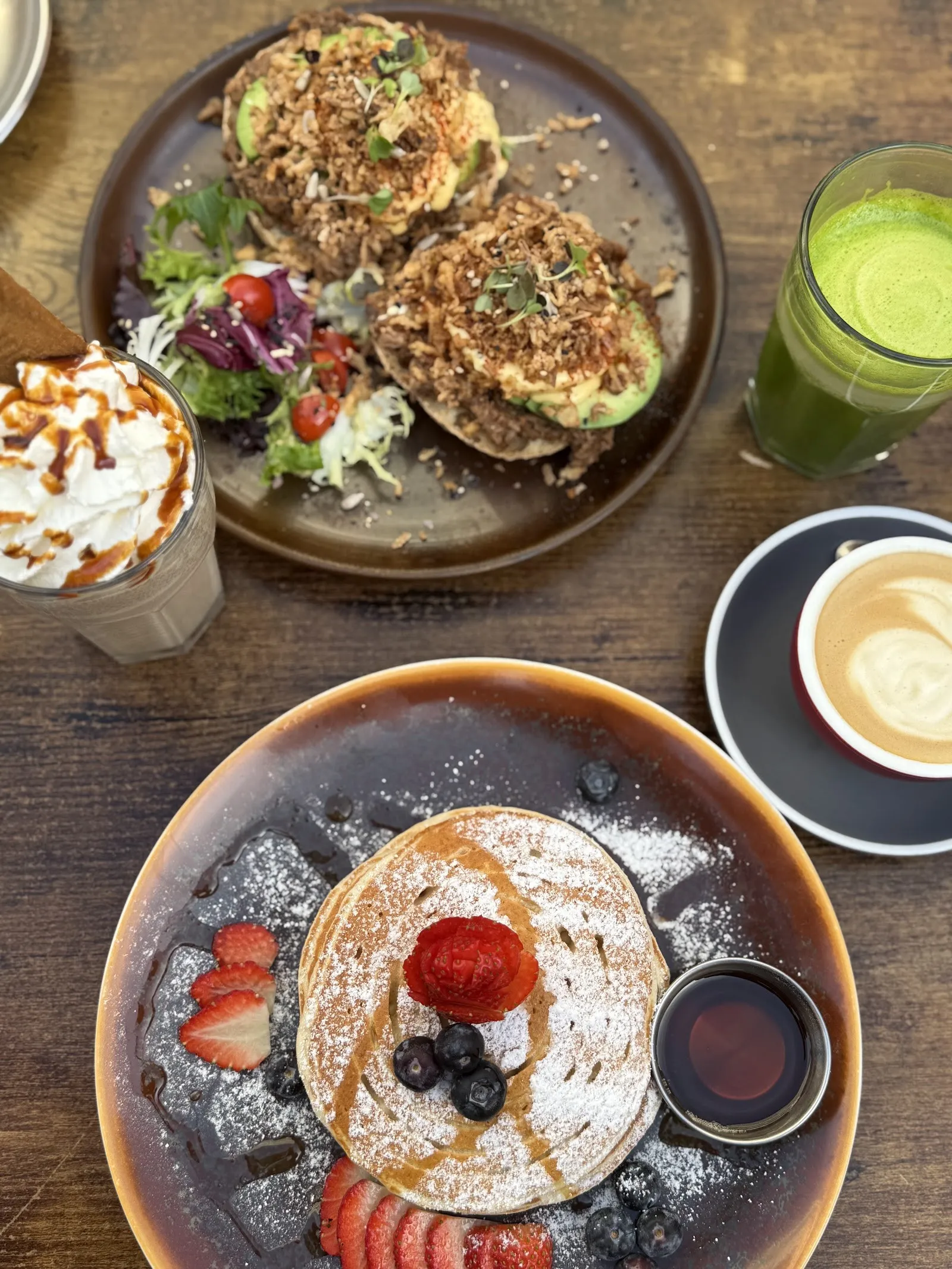 Mesa de brunch: pancakes con fruta, tostadas, un latte, un batido verde y un café con nata